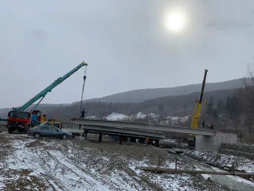 Două automacarale manevrând o grindă lungă de beton, cu munți acoperiți de zăpadă în fundal.