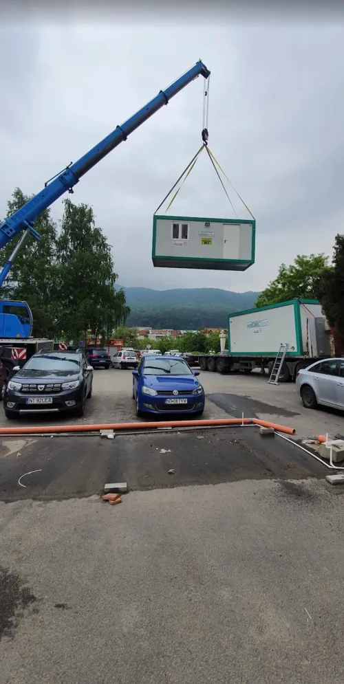 Automacara ridicând o cabină modulară verde, deasupra unei parcări cu mașini.