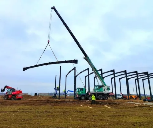 Automacara montând o grindă metalică, la structura unei hale în construcție.