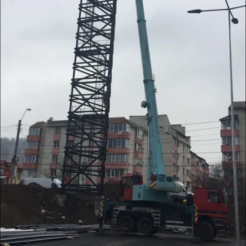 Automacara ridicând o structură metalică zăbrelită, lângă blocuri de locuințe.