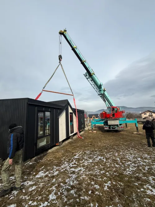 Automacara ridicând o cabină modulară neagră, la un șantier montan cu zăpadă.
