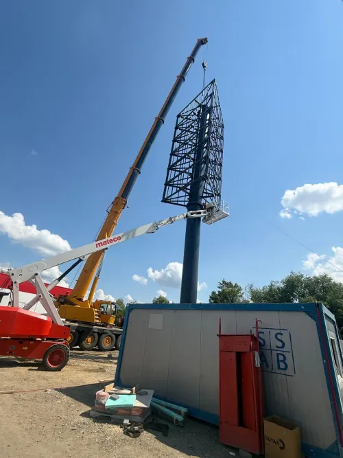Automacara și o platformă telescopică montând un panou publicitar pe stâlp, lângă o cabină modulară.